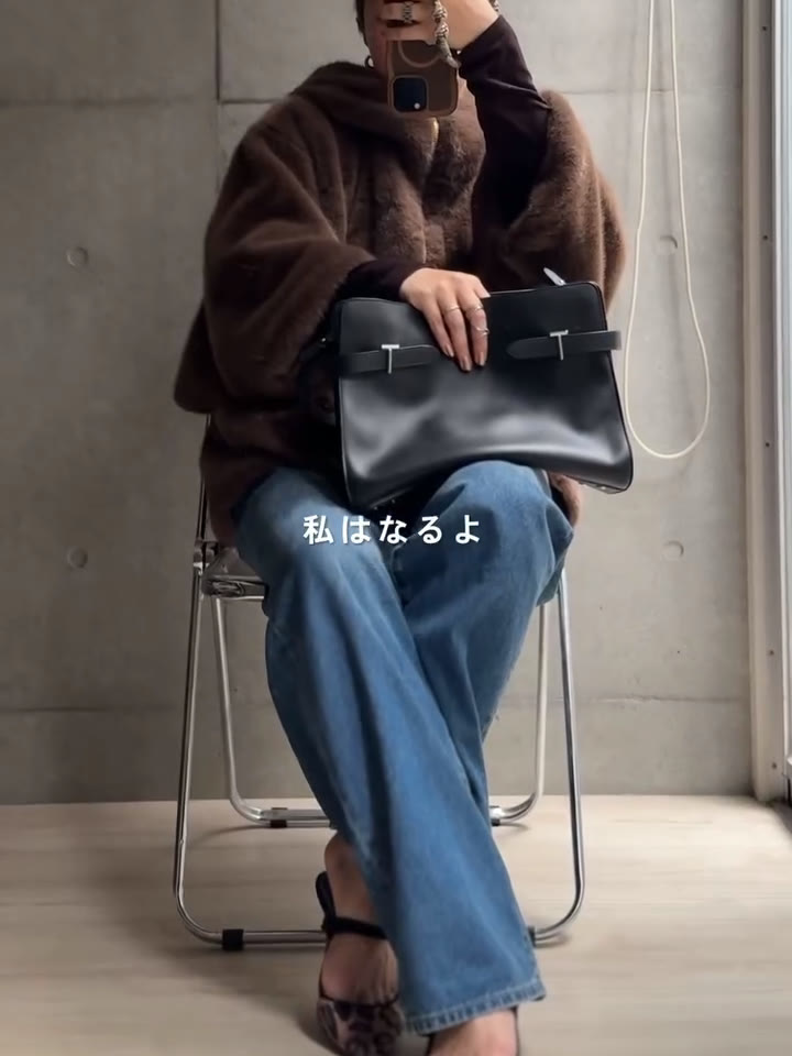 ブラウンのファーコートとデニムにLE TANNEURのEMIEショルダーバッグを合わせて椅子に座ったところ